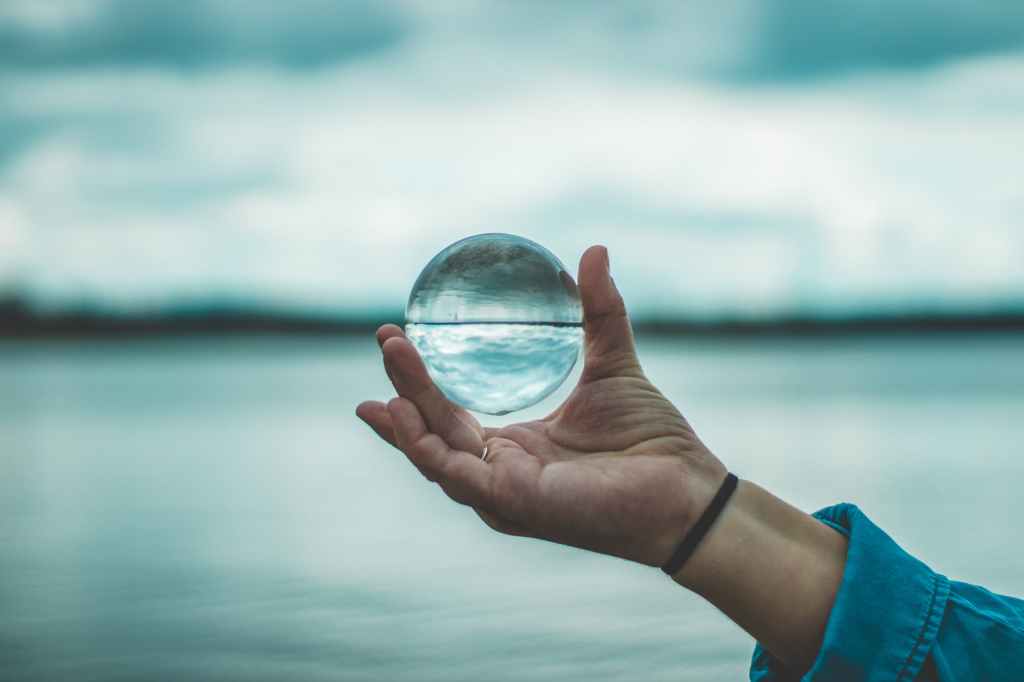 Person holding a clear sphere (lensball) with the ocean in the background.